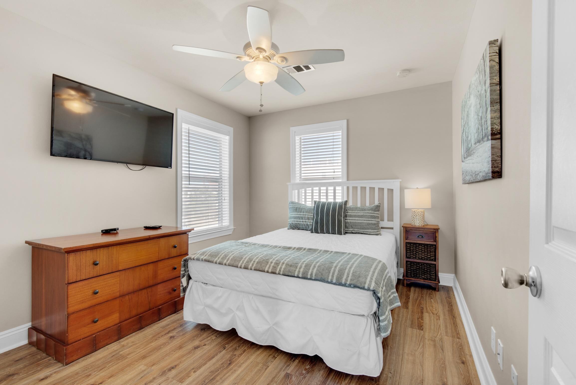 A bedroom with a bed, dresser, ceiling fan, TV, and artwork.