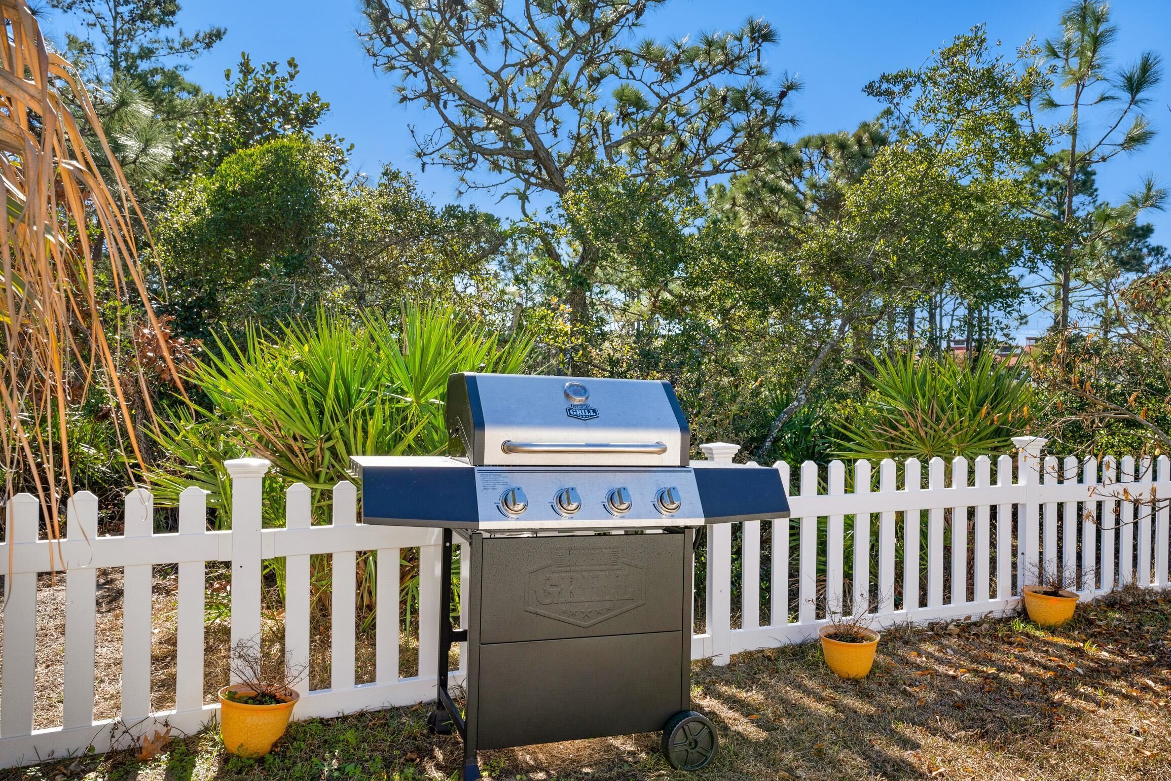 Ein tragbarer Grill mit vier Brennern, platziert in einem Garten mit weißem Zaun und Topfpflanzen.