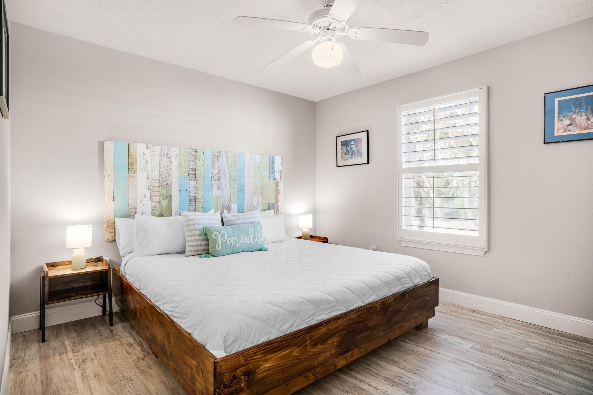 Une chambre à coucher avec un lit en bois, un ventilateur de plafond, une fenêtre avec des stores et un tableau accroché au mur.