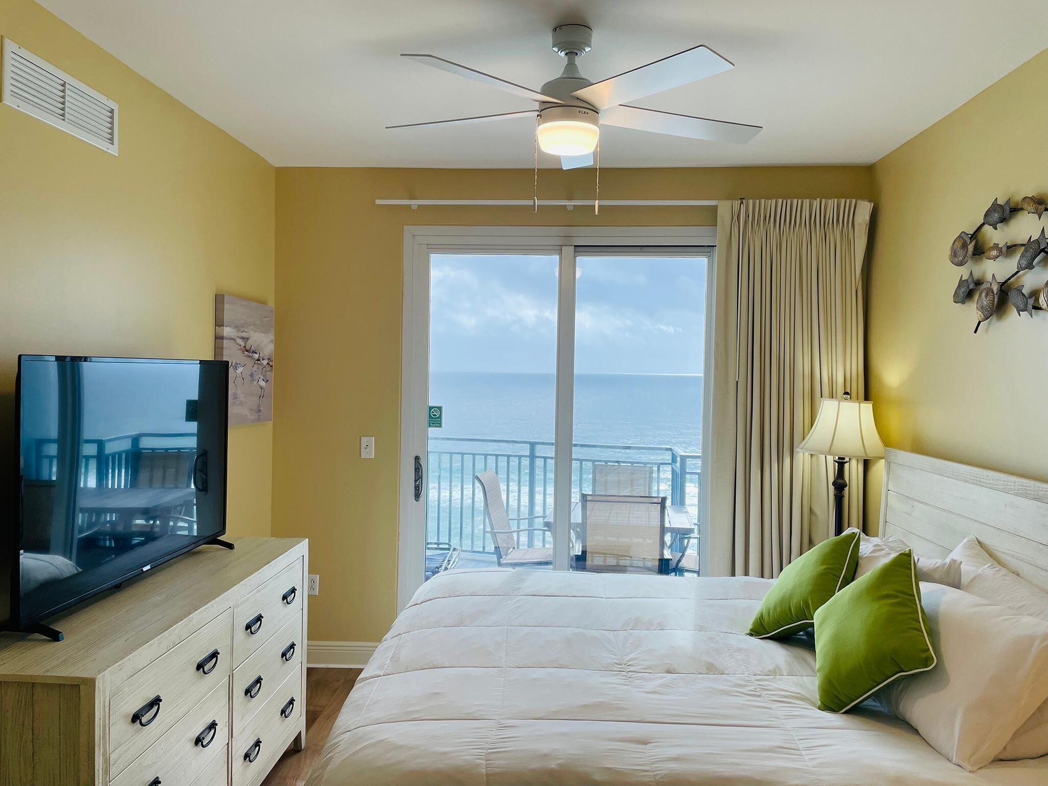 Une chambre d’hôtel équipée d’un lit, d’une télévision, d’un ventilateur de plafond, d’un balcon avec vue sur la mer et d’une lampe.
