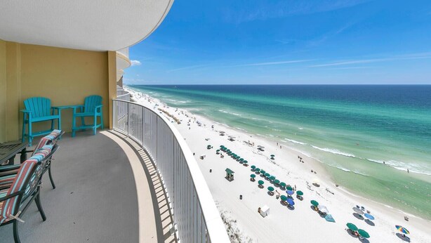 A balcony with turquoise chairs and a table, overlooking a beach with umbrellas and people.