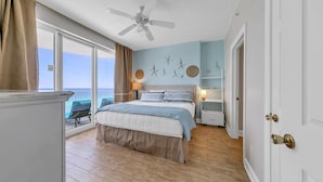 A bedroom with a bed, a ceiling fan, a nightstand, a window with curtains, and a view of the ocean.