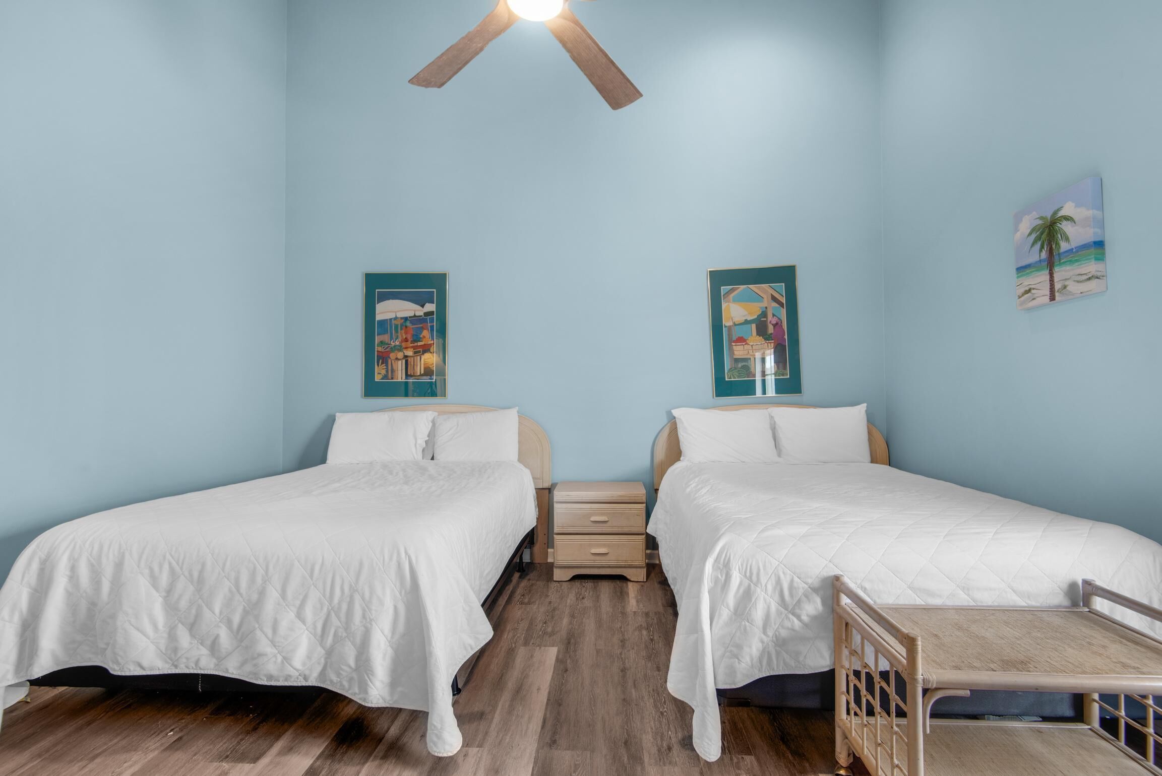 Une chambre avec deux lits, un plancher en bois, un ventilateur de plafond et deux tableaux accrochés au mur.