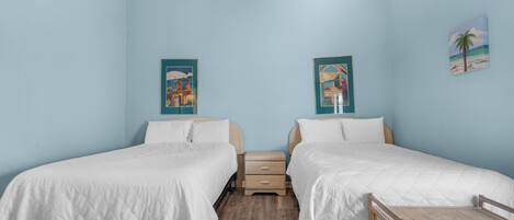 Une chambre avec deux lits, un plancher en bois, un ventilateur de plafond et deux tableaux accrochés au mur.