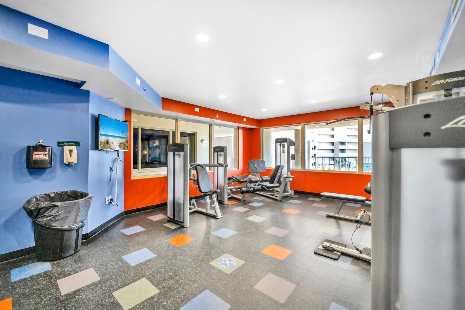 Un gimnasio con equipo de ejercicio, un bote de basura y una ventana con vistas a edificios.