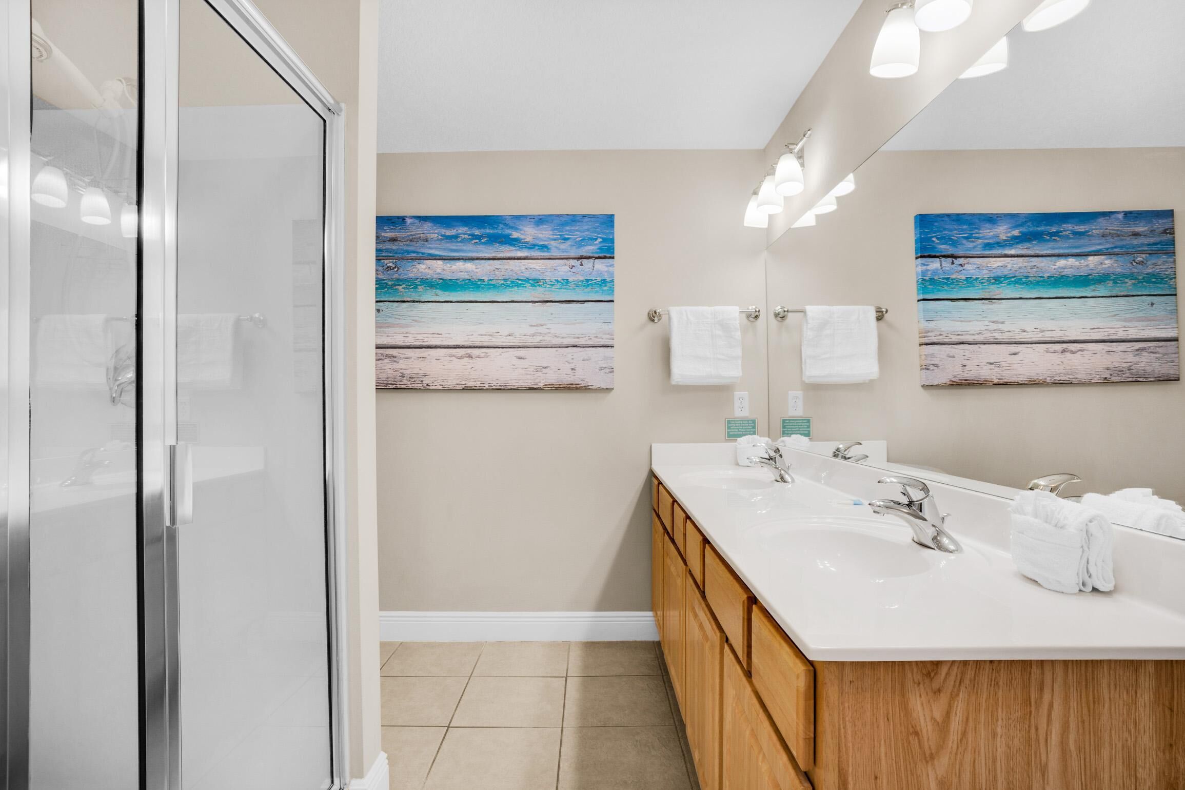 A modern bathroom with a glass shower enclosure, a white sink vanity, and two beach-themed paintings on the wall.