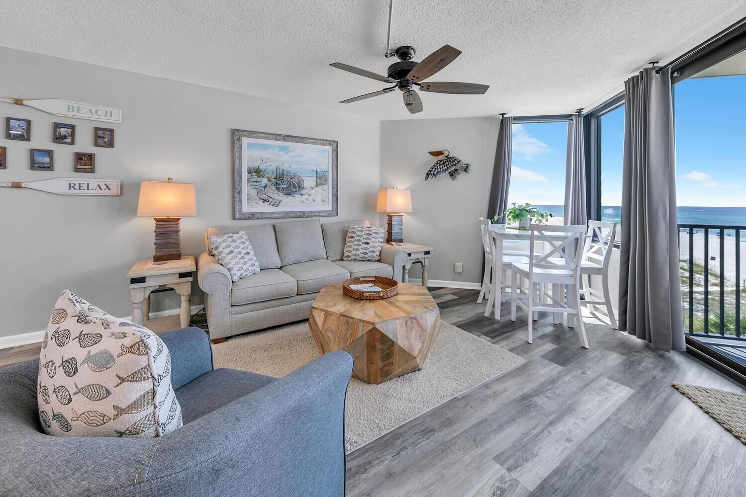 A modern living room with a sofa, two lamps, a coffee table, and a dining area with chairs. There is a ceiling fan, a painting on the wall, and a view of the ocean through large windows.