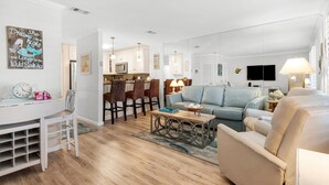 Un salon spacieux avec un canapé bleu clair, un fauteuil beige et une table basse en bois. On y trouve également un coin repas avec des chaises en osier et une table blanche. La cuisine, dotée d’appareils électroménagers modernes, est visible au fond.