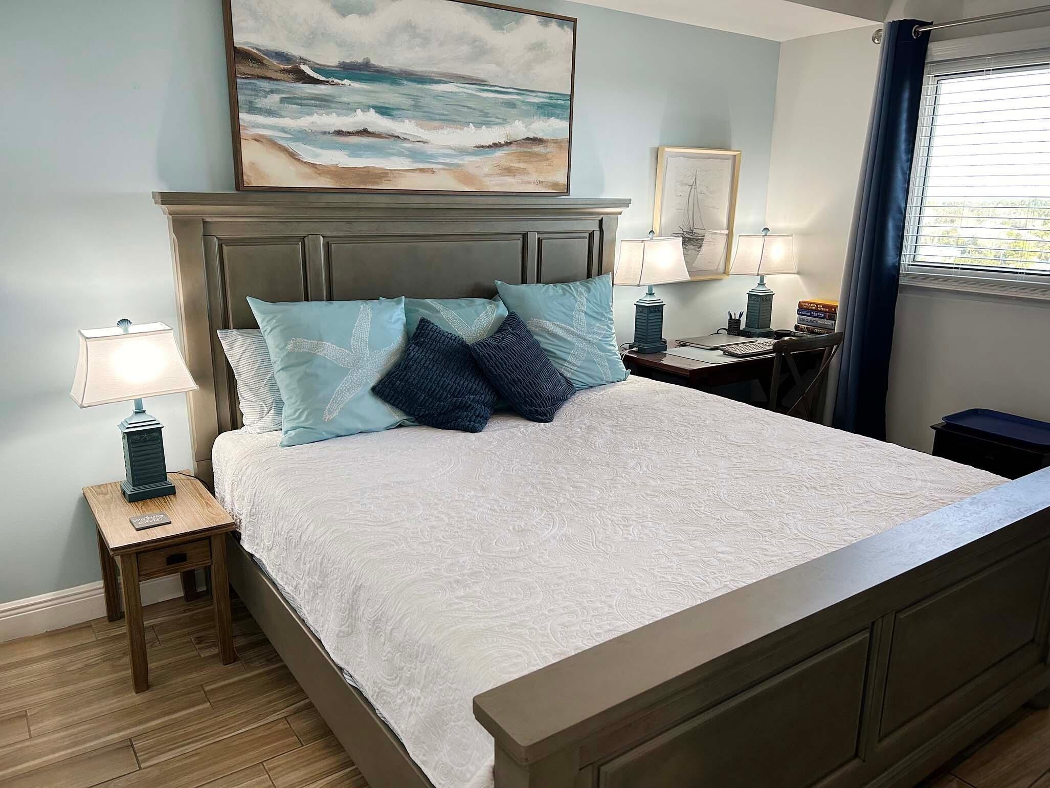 A bedroom with a bed, nightstands, a lamp, a painting, and a window with curtains.