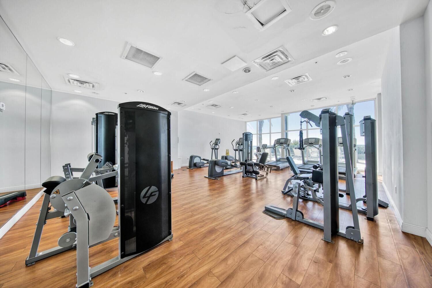 A modern gym with various exercise equipment, large windows, and a wooden floor.