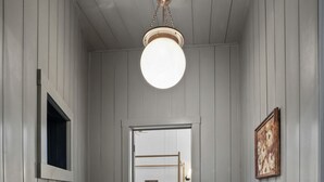 Un pasillo con piso de madera, un candelabro en el techo, una puerta que da a un dormitorio y una pequeña mesa con una planta.