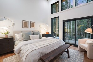 Ein Schlafzimmer mit einem großen Bett, zwei Nachttischen, einer Bank, einem Sessel und großen Fenstern mit Blick auf einen Wald.