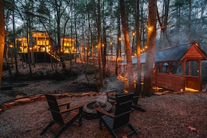 Un chalet en forêt, doté d’un foyer et d’un espace détente, entouré d’arbres et illuminé de guirlandes lumineuses.