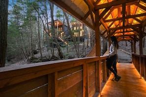 A wooden deck with a person standing, overlooking a forested area and a building with lights.