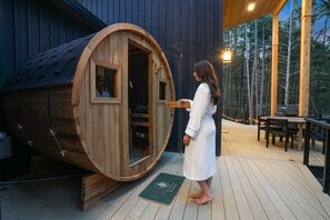 A wooden sauna with a door open, a wooden deck with a table and chairs, and a forested background.