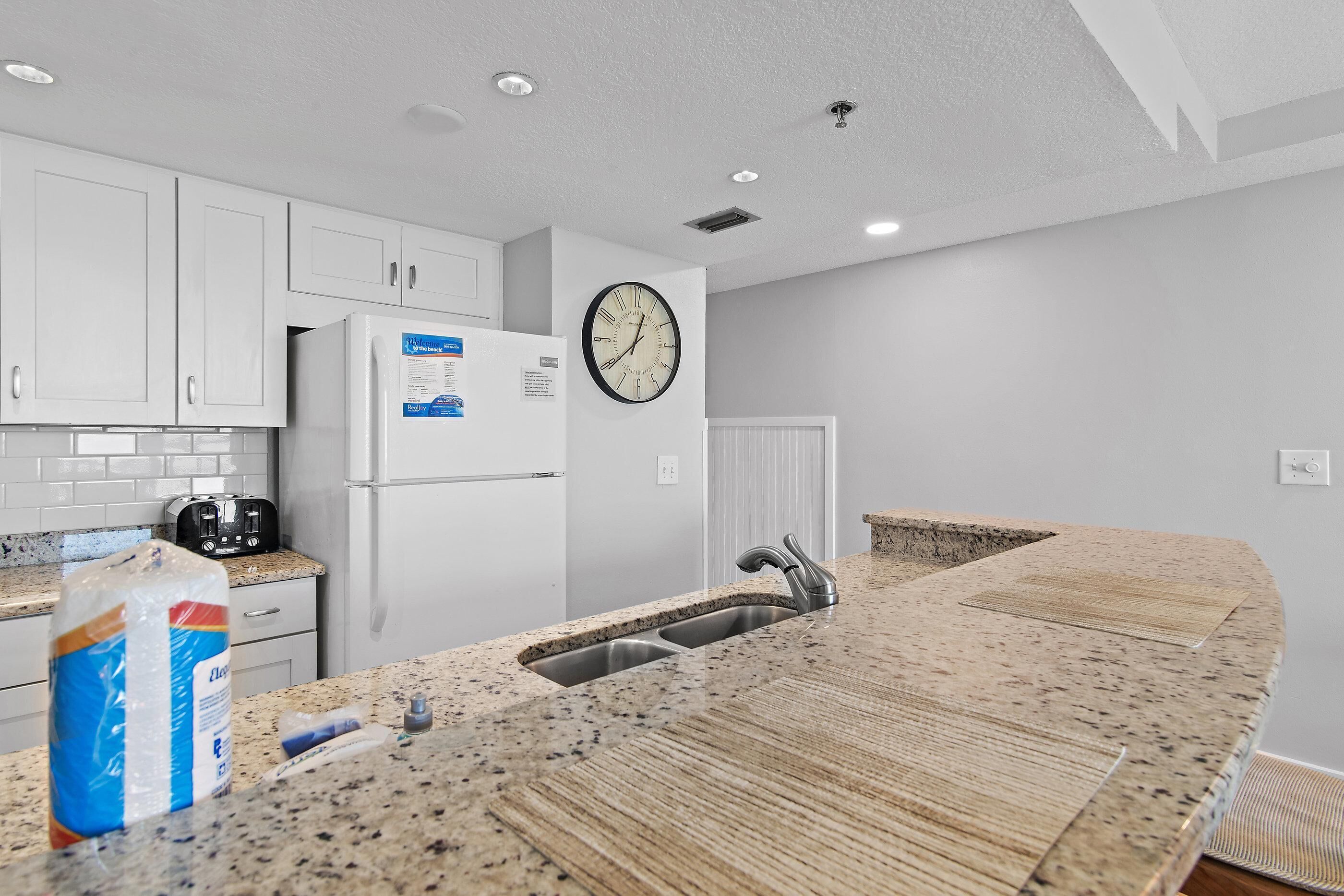 A kitchen with white cabinets, a granite countertop, a stainless steel sink, and a clock on the wall.