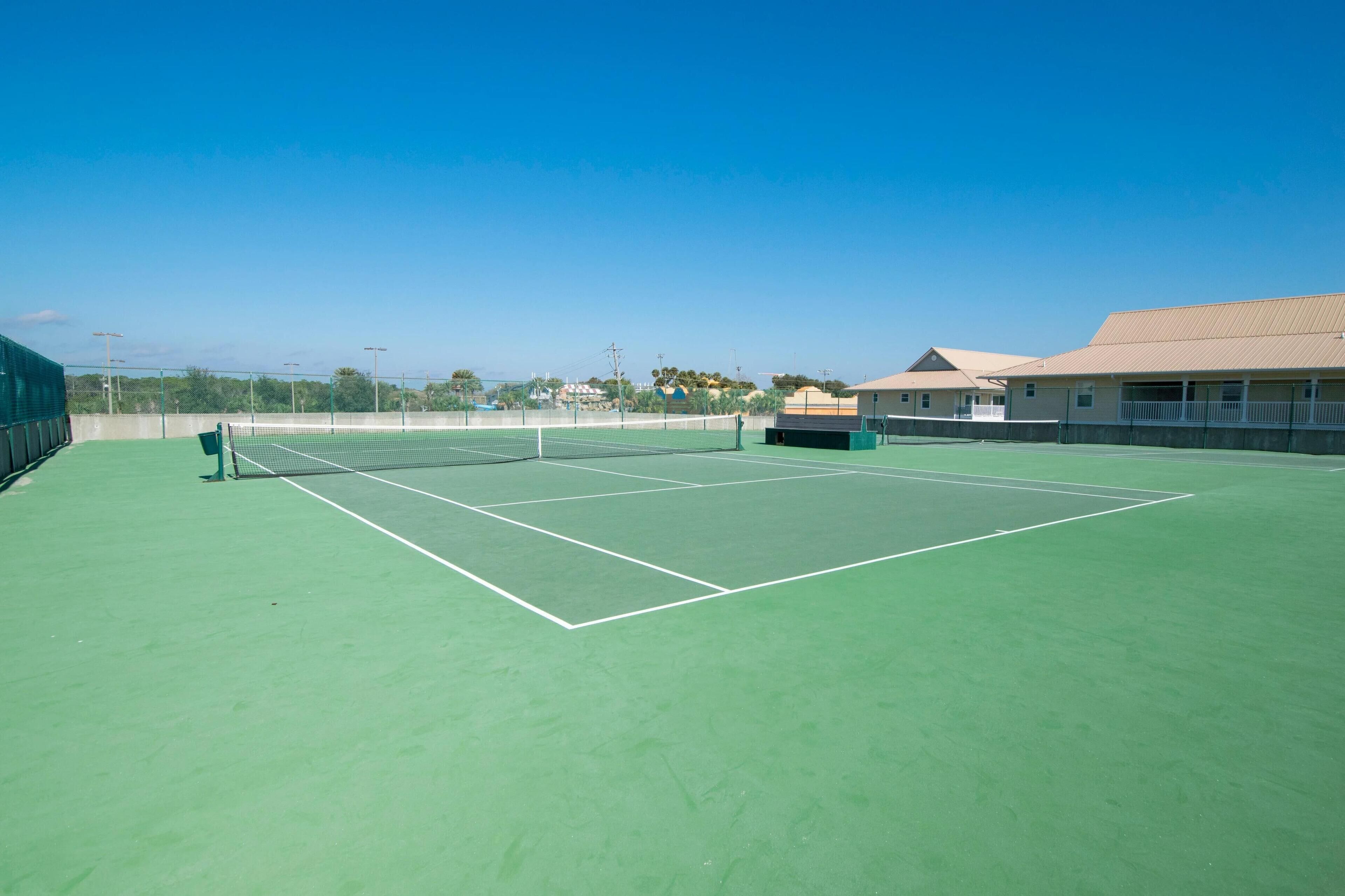 A tennis court with a net, surrounded by a green surface and a building with a beige roof.