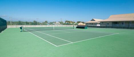 A tennis court with a net, surrounded by a green surface and a building with a beige roof.