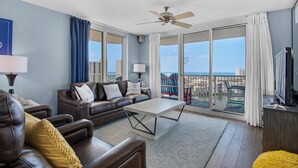 A modern living room with a brown leather sofa, a white coffee table, and a large window offering a view of the city and the sea. - Pelican Beach Terrace 807 (Destin)