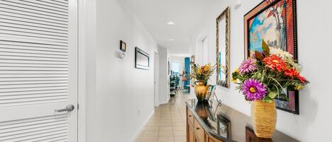 A hallway with a white door, a wooden cabinet with a vase of flowers, and framed artwork on the wall.
