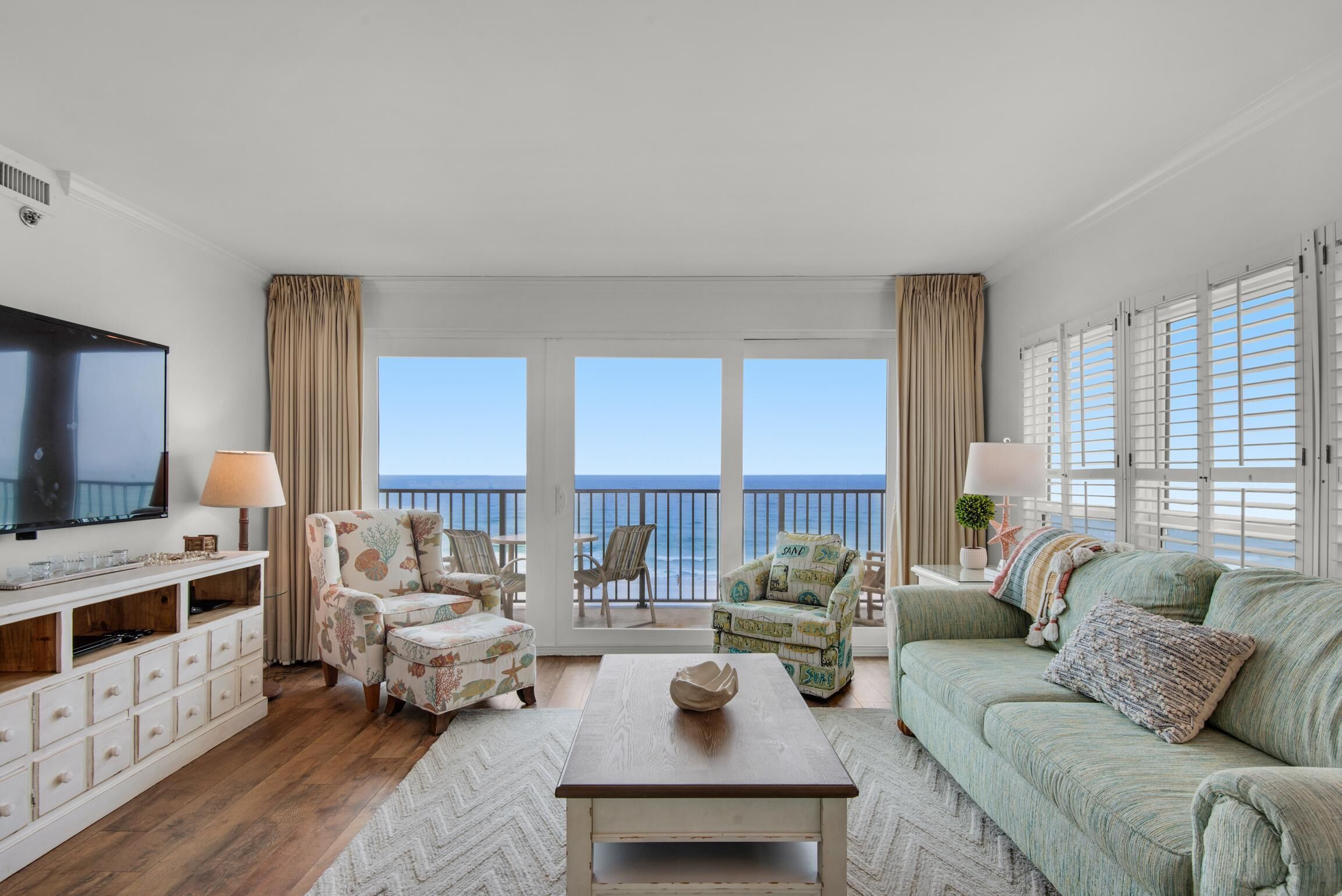 A spacious living room with a large flat-screen TV, comfortable seating, a coffee table, and a view of the ocean through large windows.