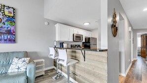 A modern kitchen with white cabinets, a stainless steel refrigerator, and a microwave. There is a bar area with two white bar stools. The room has wooden flooring and a light gray wall.