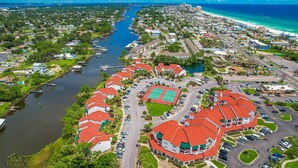 An aerial view of a coastal residential area with a river, a tennis court, and a large building with a red roof.