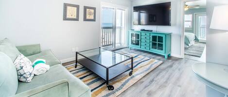 A living room with a sofa, a glass coffee table, a teal cabinet, a flat-screen TV, and a ceiling fan.