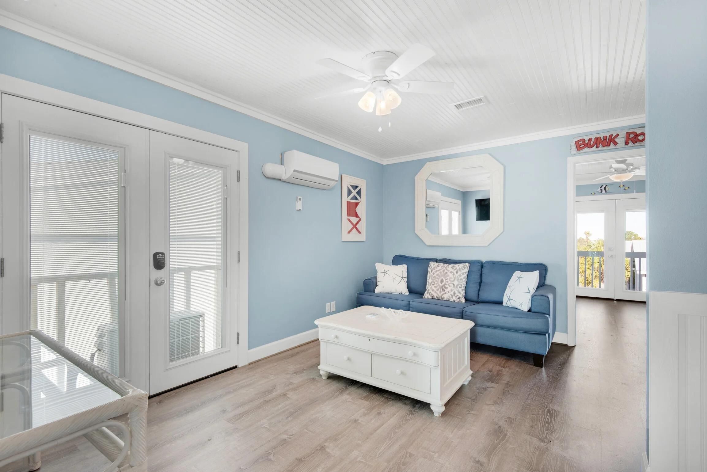 Una sala de estar con pared azul, techo blanco y ventilador de techo. Hay una mesa de centro blanca, un sofá azul con cojines estampados y una mesita auxiliar blanca.