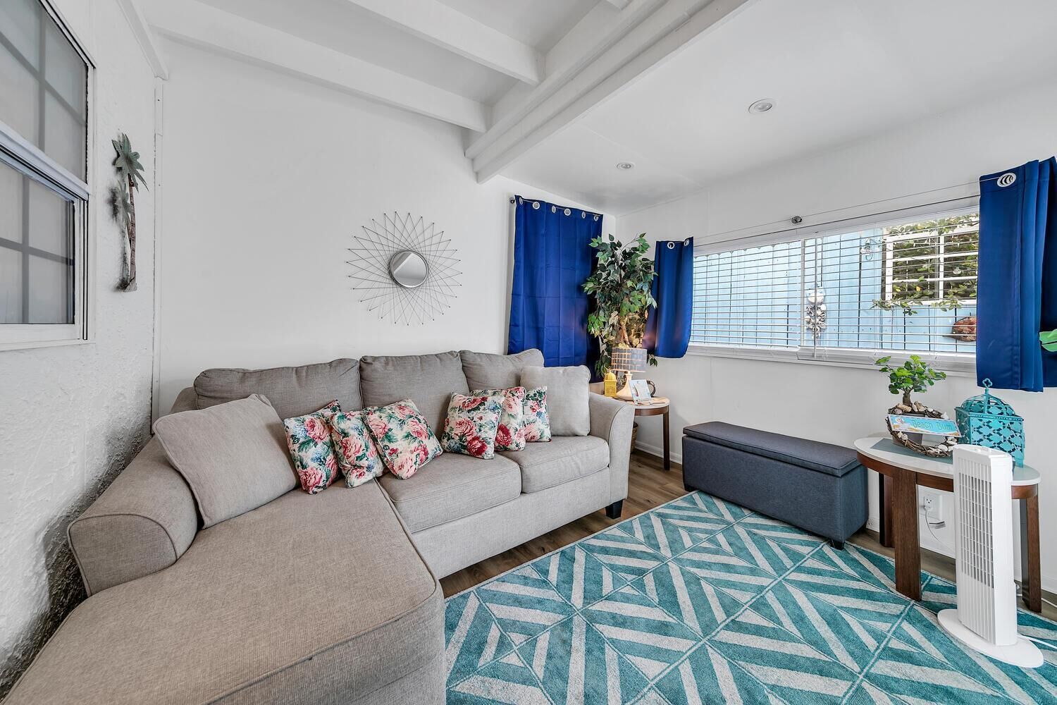 A living room with a grey sofa, floral cushions, a blue patterned rug, a small side table with a plant, and a window with blue curtains.