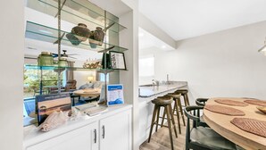 Une cuisine moderne avec une étagère en verre où sont exposés des objets décoratifs, une table de salle à manger ronde avec des tabourets, et une vue sur l’extérieur.
