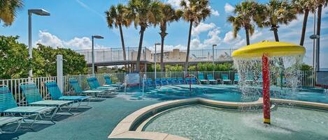 A swimming pool with blue lounge chairs, a water feature, palm trees, and a covered walkway.