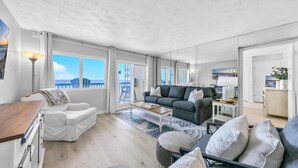 En rummelig opholdsstue med en grå sofa, hvide lænestole og et sofabord. Der er en balkon med udsigt over byens skyline.