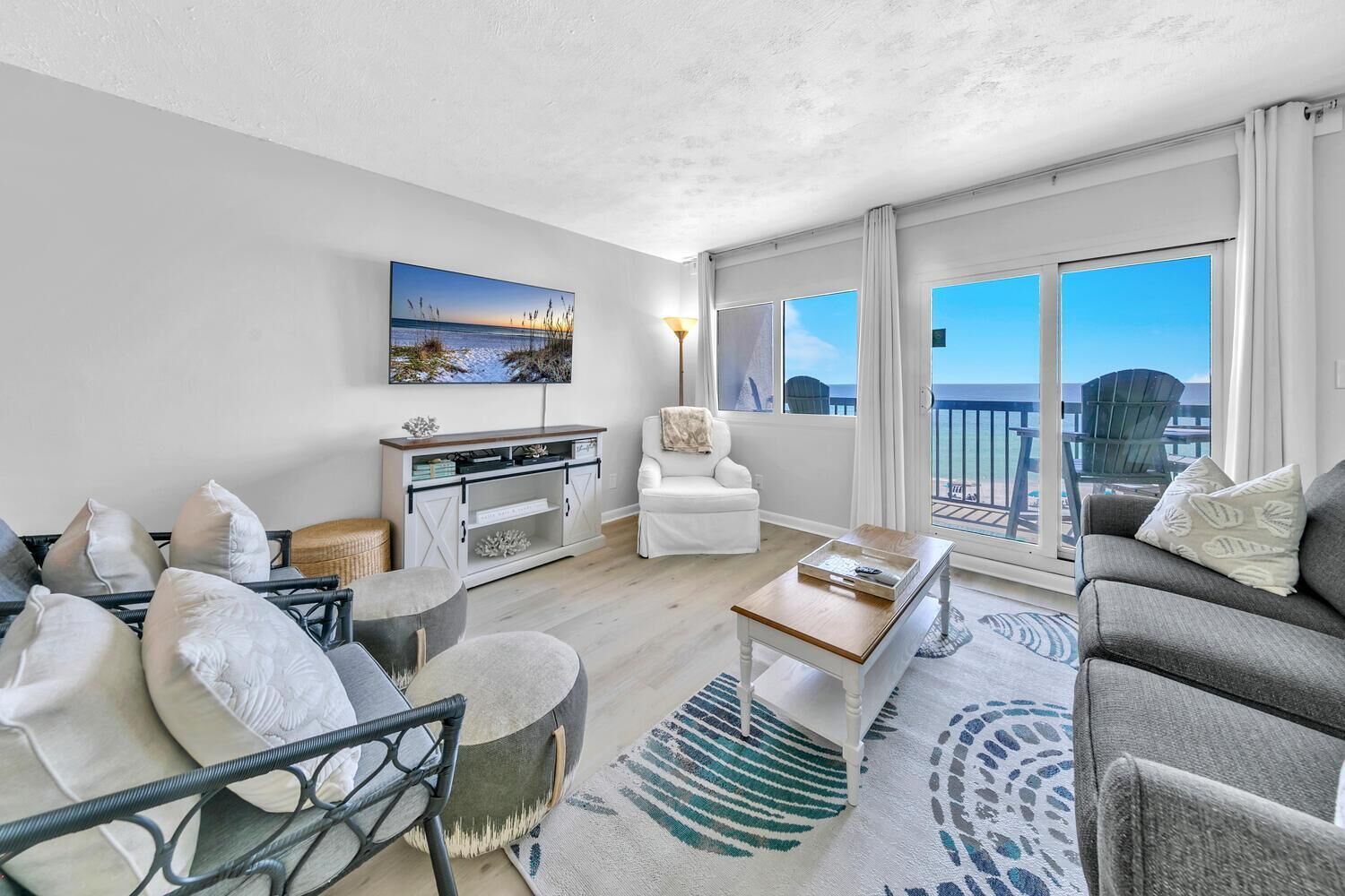 A spacious living room with a white sofa, a grey sofa, a coffee table, and a wicker chair. There is a white cabinet with decorative items and a painting of a beach scene. The room has large windows with a view of the ocean.