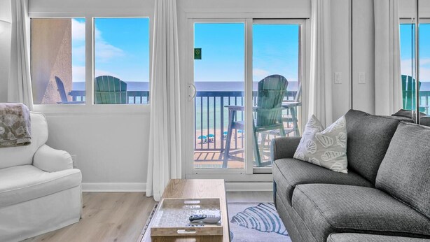 Et værelse med en hvid sofa, en grå sofa og et træsofabord. To vinduer med hvide gardiner, samt udsigt til stranden og stole udenfor.