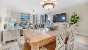 A modern living room with a wooden dining table, white chairs, a chandelier, a flat-screen TV, a ceiling fan, and a large window with a view. - Mainsail 234 (Miramar Beach)