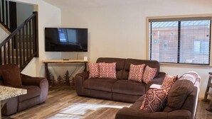 A living room with a brown sofa set, a flat-screen TV mounted on the wall, a wooden staircase, and a window with blinds.