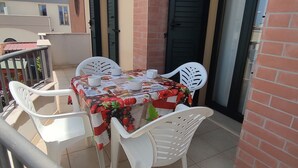 Un balcon avec une table dressée pour deux personnes, des chaises en plastique blanches et une nappe à motifs floraux.