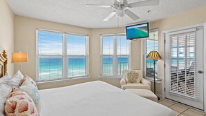 A bedroom with a bed, a ceiling fan, a TV, and a view of the ocean through large windows. - The Palms #601 (Fort Walton Beach)
