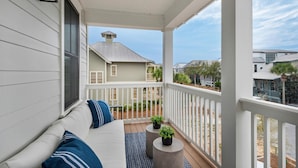 A covered porch with a white railing, a white sofa with blue cushions, and two potted plants on round stools.
