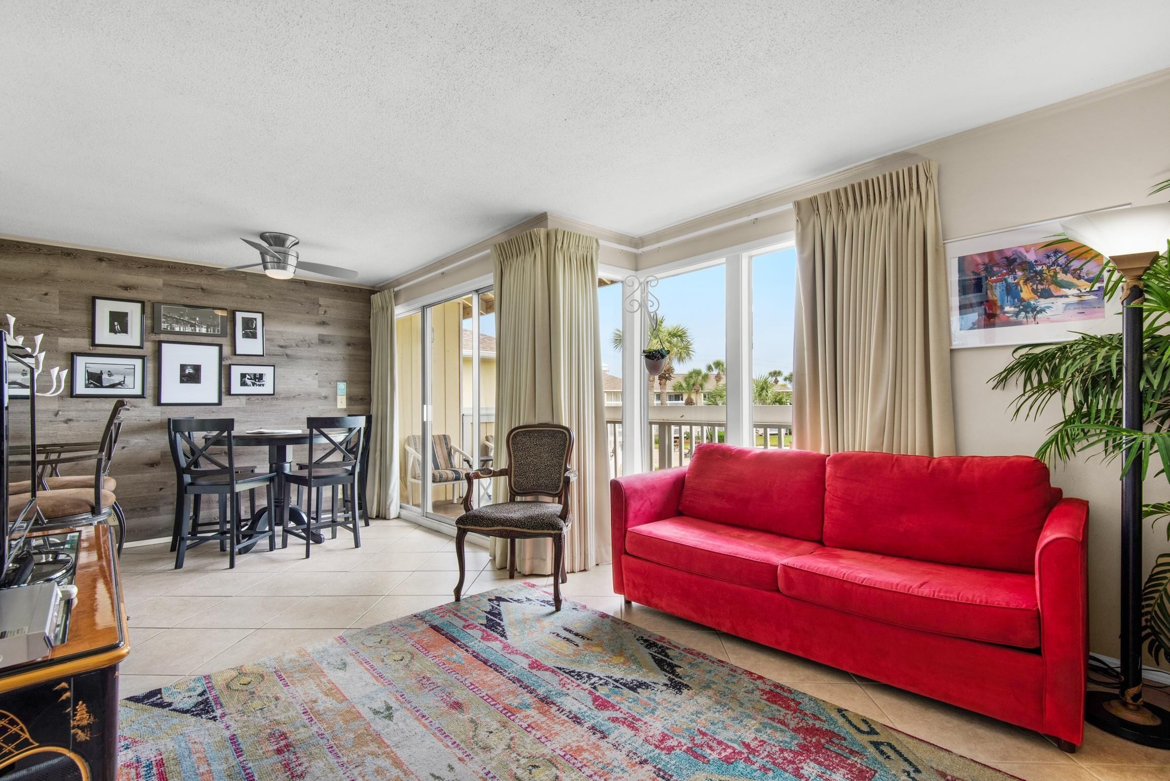 A living room with a red sofa, a patterned rug, a dining area with chairs, a ceiling fan, and a large window with curtains.