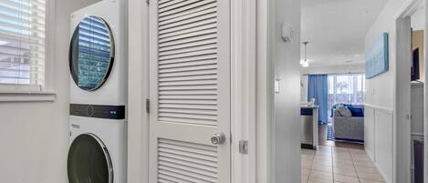 Une buanderie équipée d’un lave-linge et d’un sèche-linge à chargement frontal, d’une porte à panneaux ajourés et d’un couloir menant à un espace de vie comprenant un canapé et un coin repas.