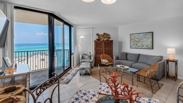 Een ruime woonkamer met een balkon dat uitkijkt op het strand, een grijze bank, een houten kast en een eethoek met een tafel en stoelen.
