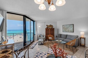 A spacious living room with a balcony overlooking the beach, a grey sofa, a wooden cabinet, and a dining area with a table and chairs. - Sunbird 609e (Panama City Beach)