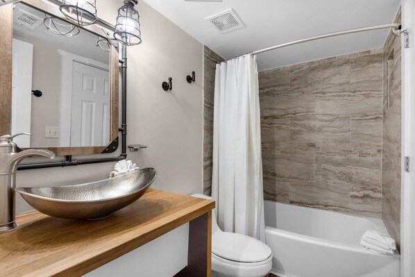 A bathroom with a wooden vanity, a vessel sink, a white toilet, and a shower with a stone wall.