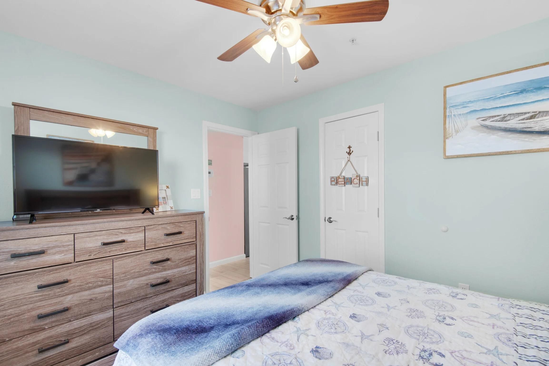 A bedroom with a wooden dresser, a bed with a blue and white patterned bedspread, a ceiling fan, and a painting of a boat on the wall.