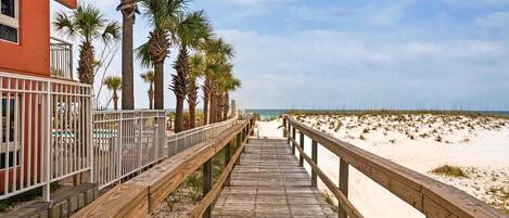 A wooden boardwalk leading to a beach with palm trees and a building on the left.
