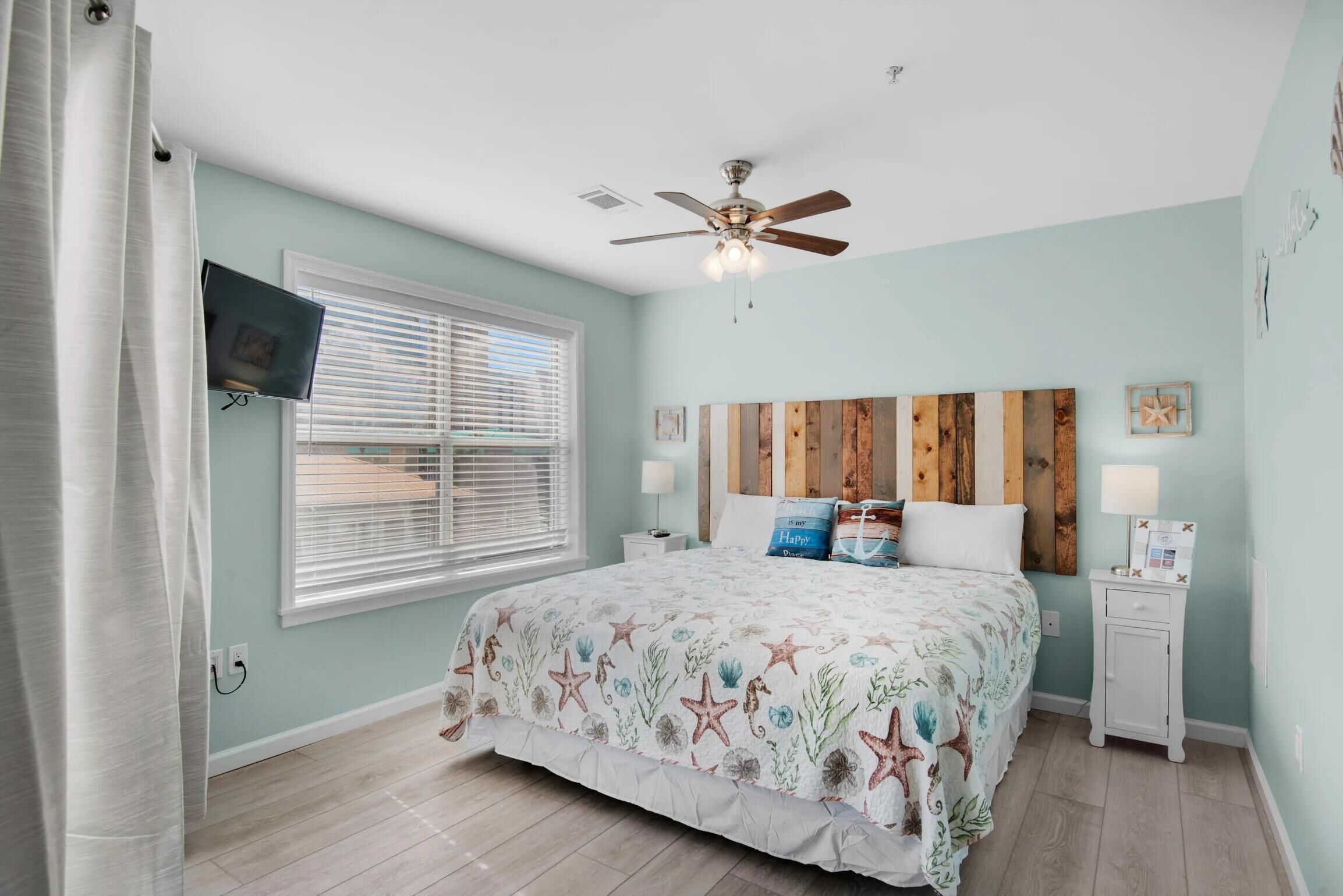 A bedroom with a bed, a ceiling fan, a TV mounted on the wall, and a window with blinds.