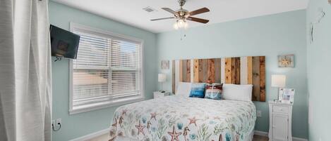 A bedroom with a bed, a ceiling fan, a TV mounted on the wall, and a window with blinds.