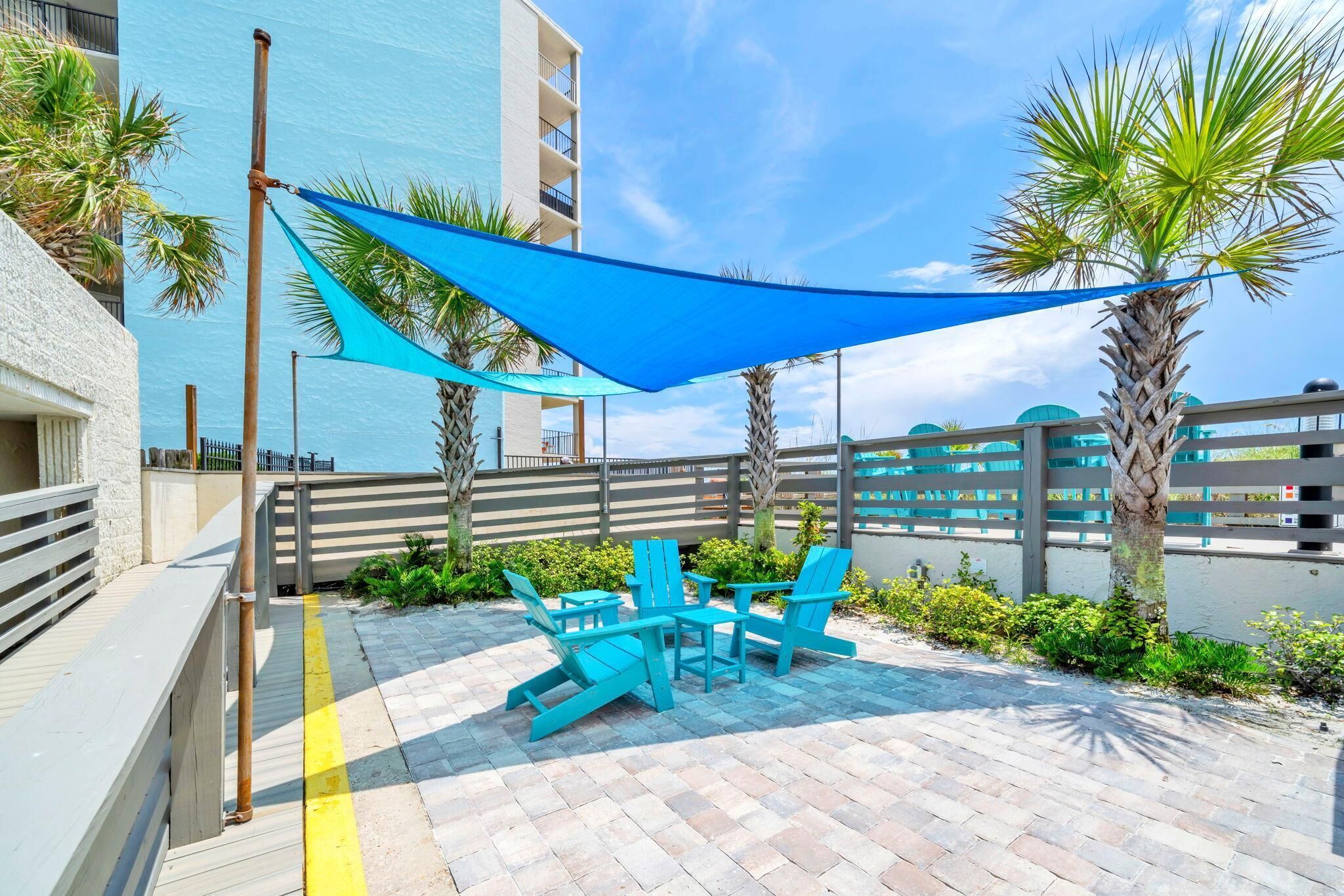Eine Terrasse im Freien mit blauen Stühlen und einem Tisch, einem blauen Sonnensegel und einem weißen Gebäude im Hintergrund.
